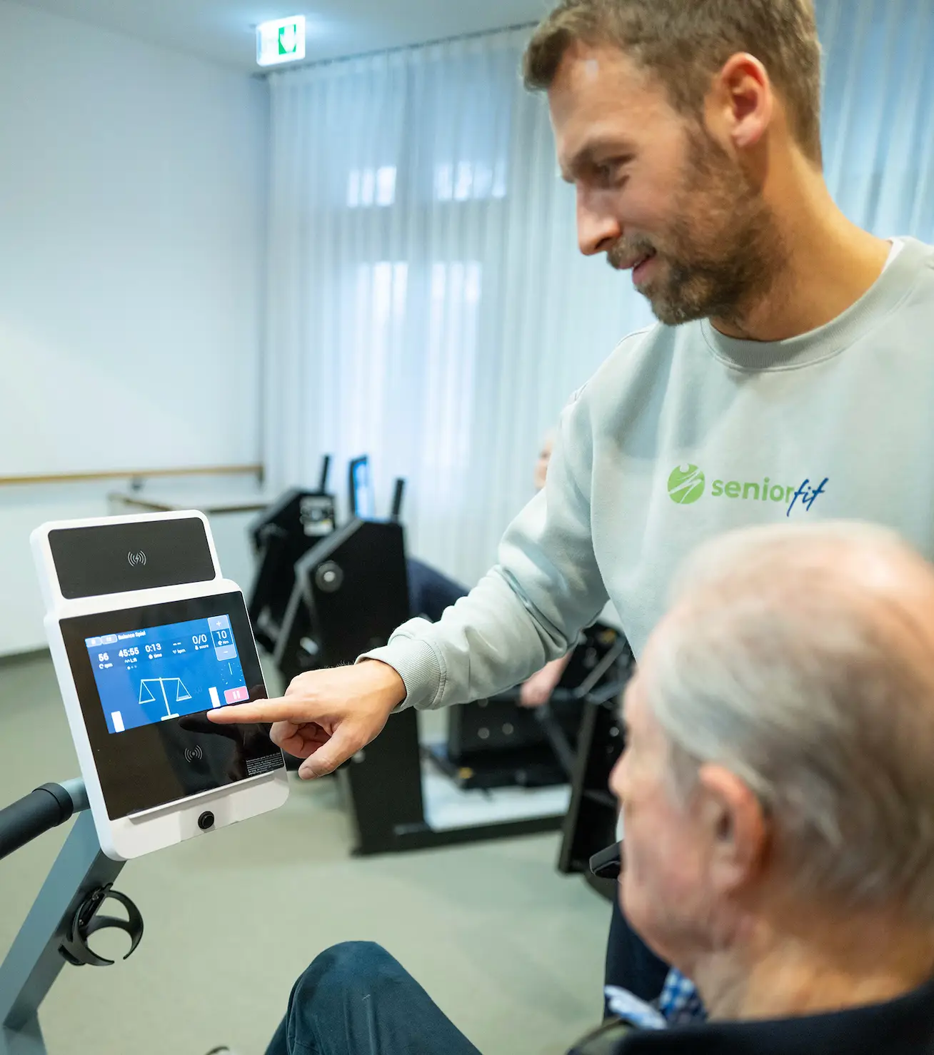 SeniorFit-Trainer erklärt einem Bewohner das Touchdisplay des Trainingsgeräts